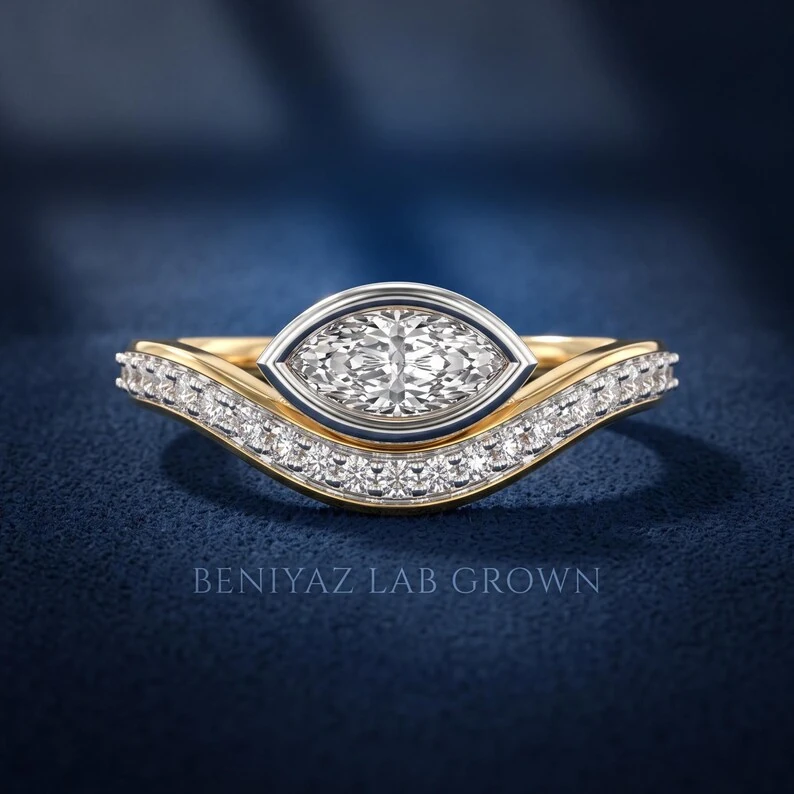 East west marquise lab grown diamond engagement ring in a white gold bezel with yellow gold curved pavé shoulders on deep navy fabric.